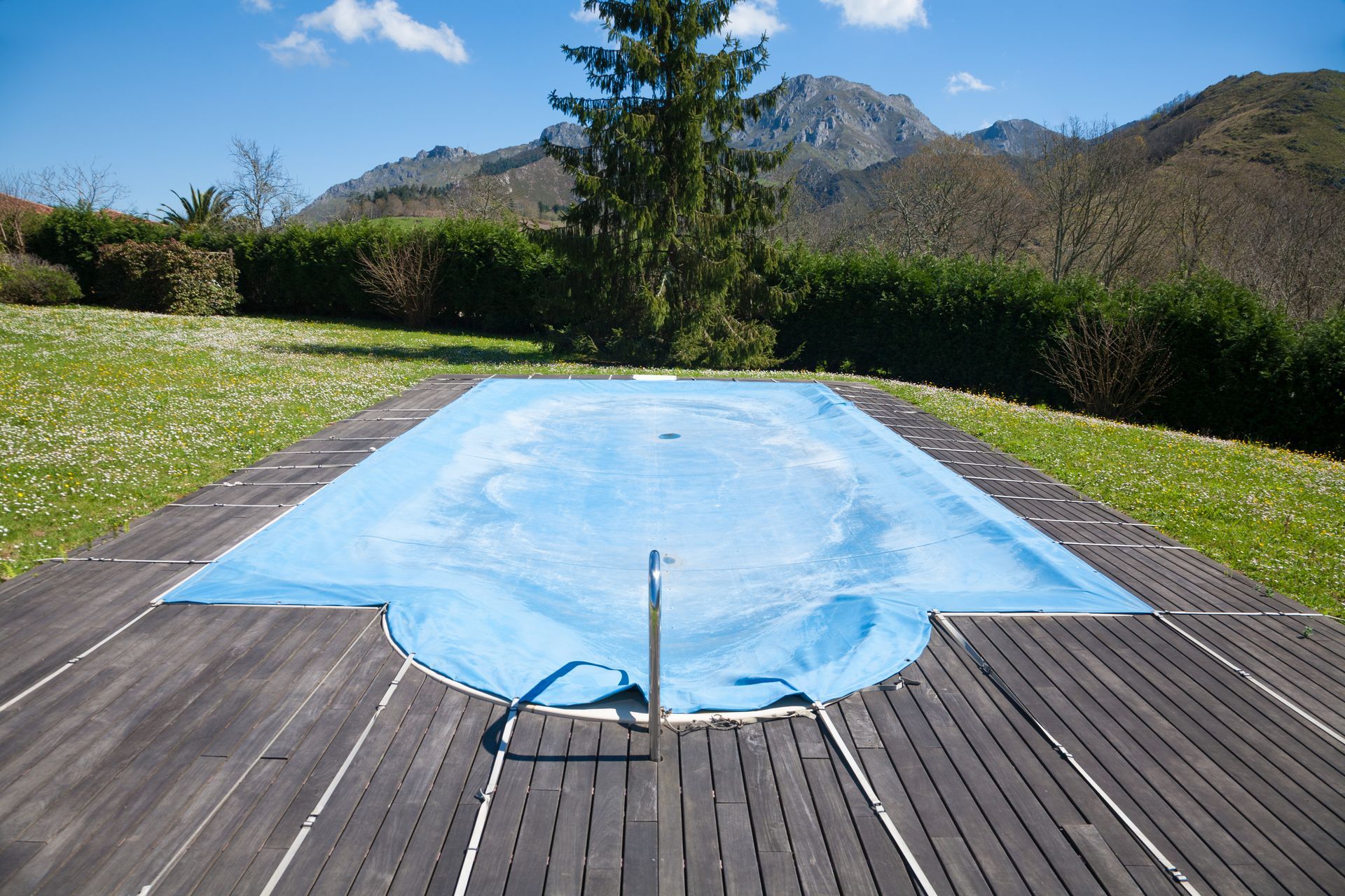 Une bâche bleue recouvre une piscine extérieure, avec des montagnes et des arbres en arrière-plan.