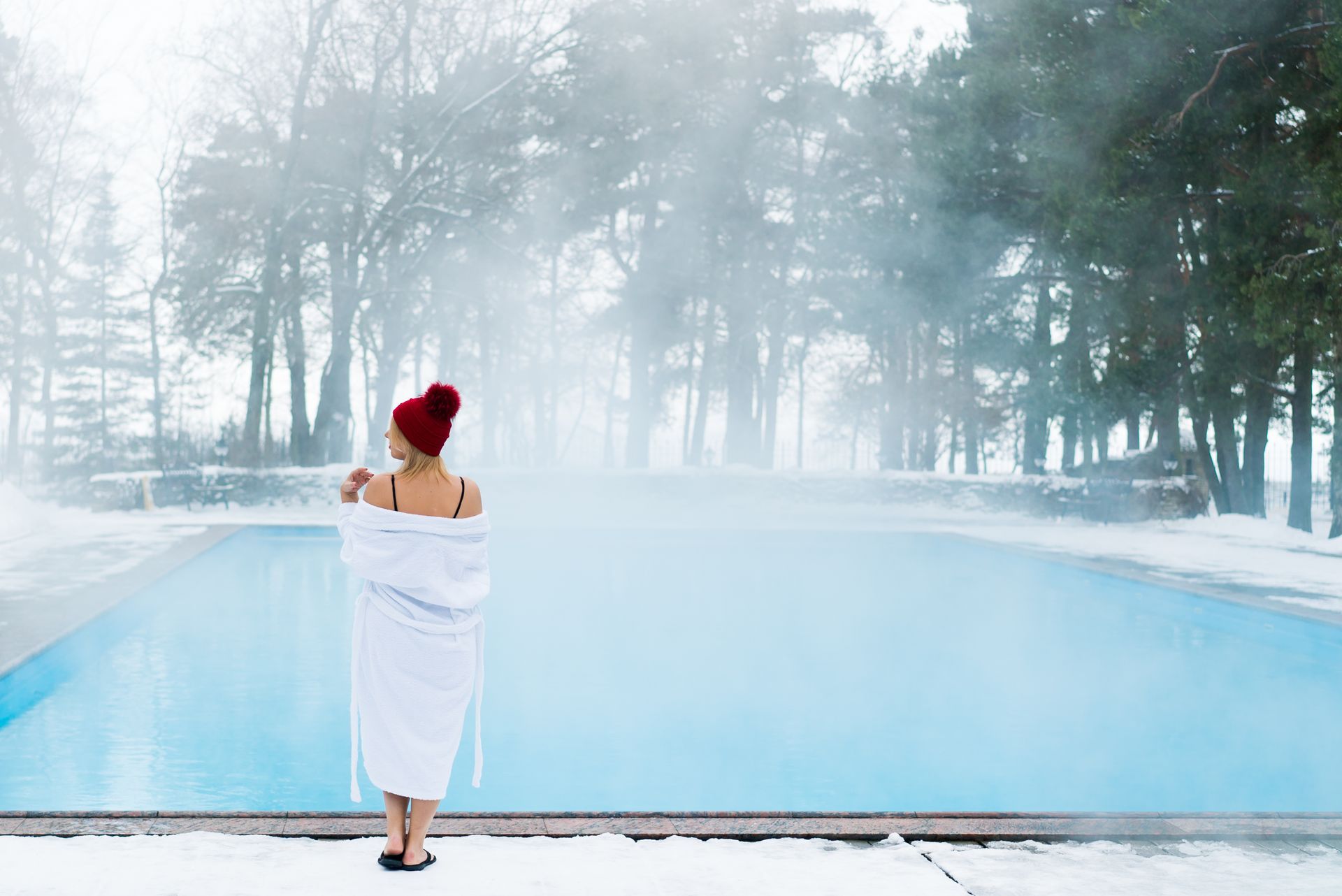 Une personne en serviette blanche se tient debout au bord d'une piscine extérieure brumeuse en hiver.