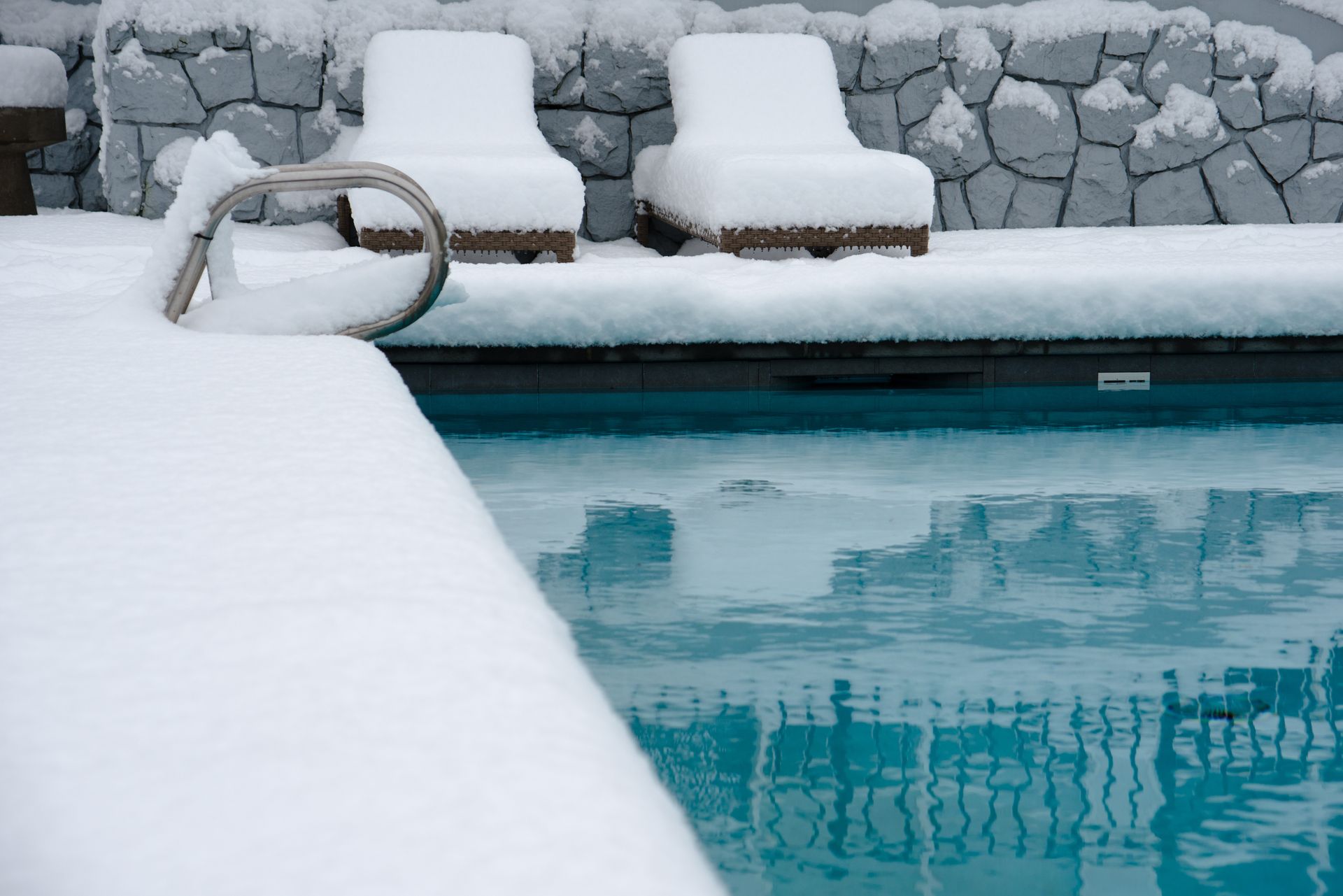 Bord de piscine enneigé avec un mur en pierre et des chaises longues couvertes de neige