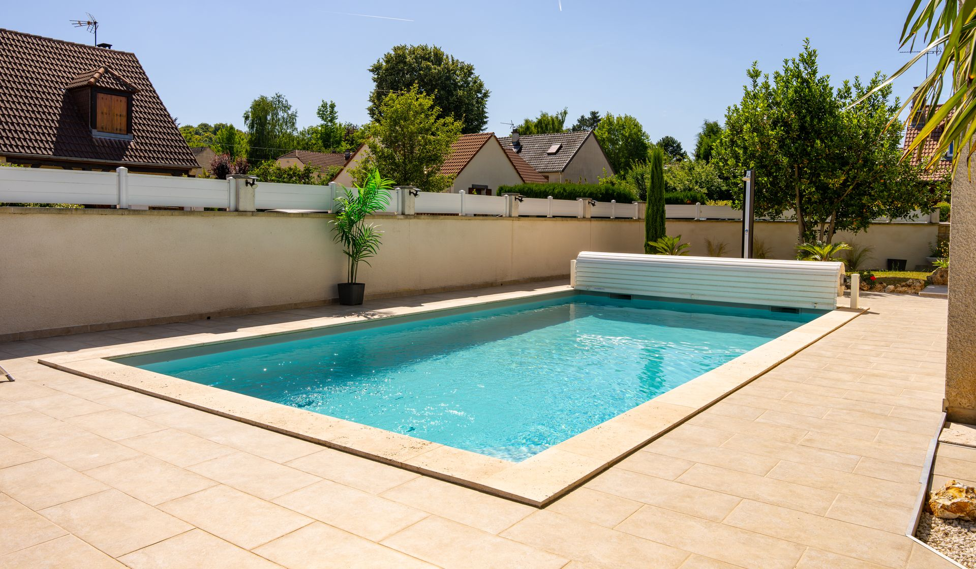 Piscine rectangulaire sur une terrasse carrelée dans le jardin, entourée d'un mur blanc sous un ciel ensoleillé.