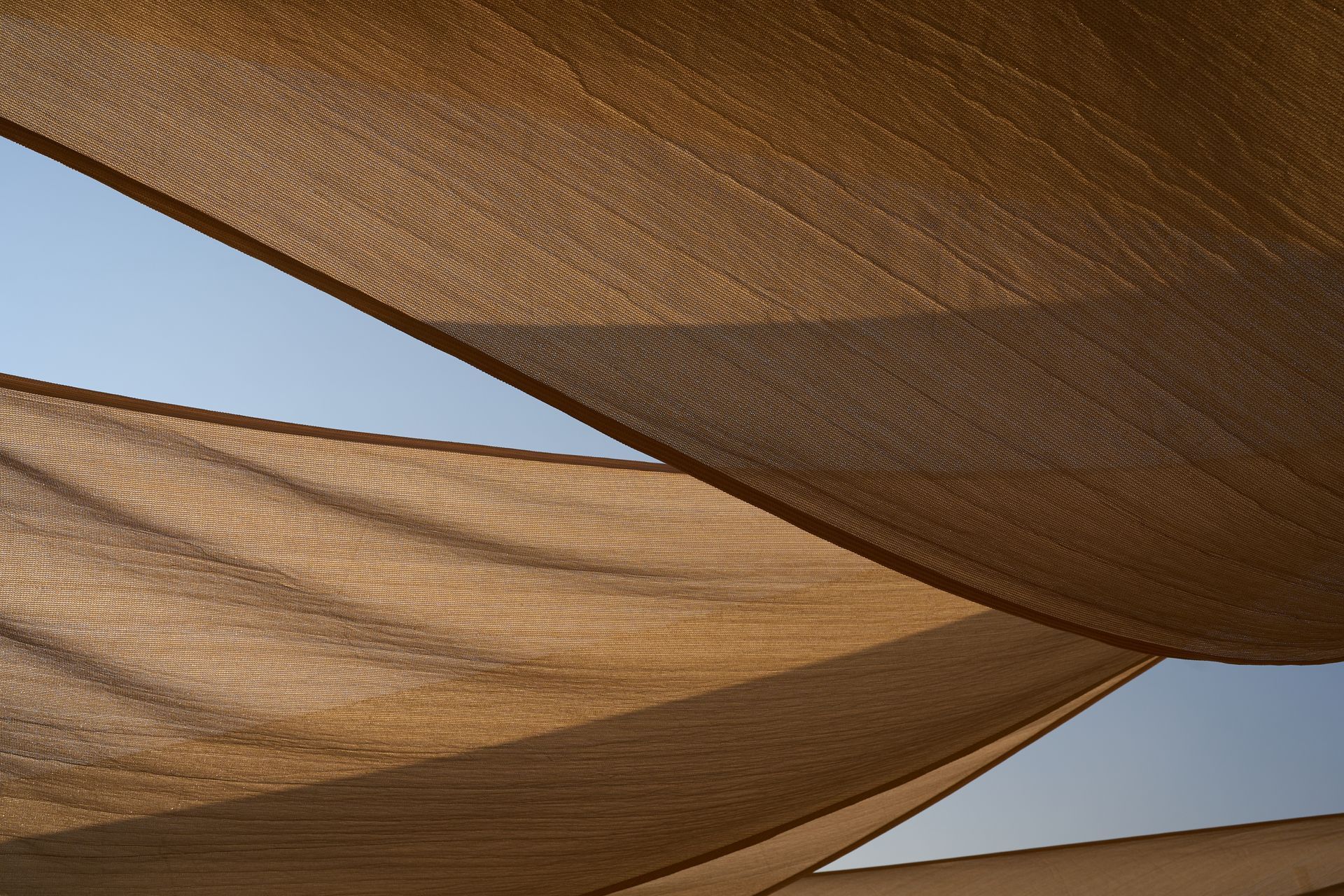 Des voiles d'ombrage tissées couleur beige s'étendent sur un ciel bleu, formant des motifs géométriques entrecroisés.