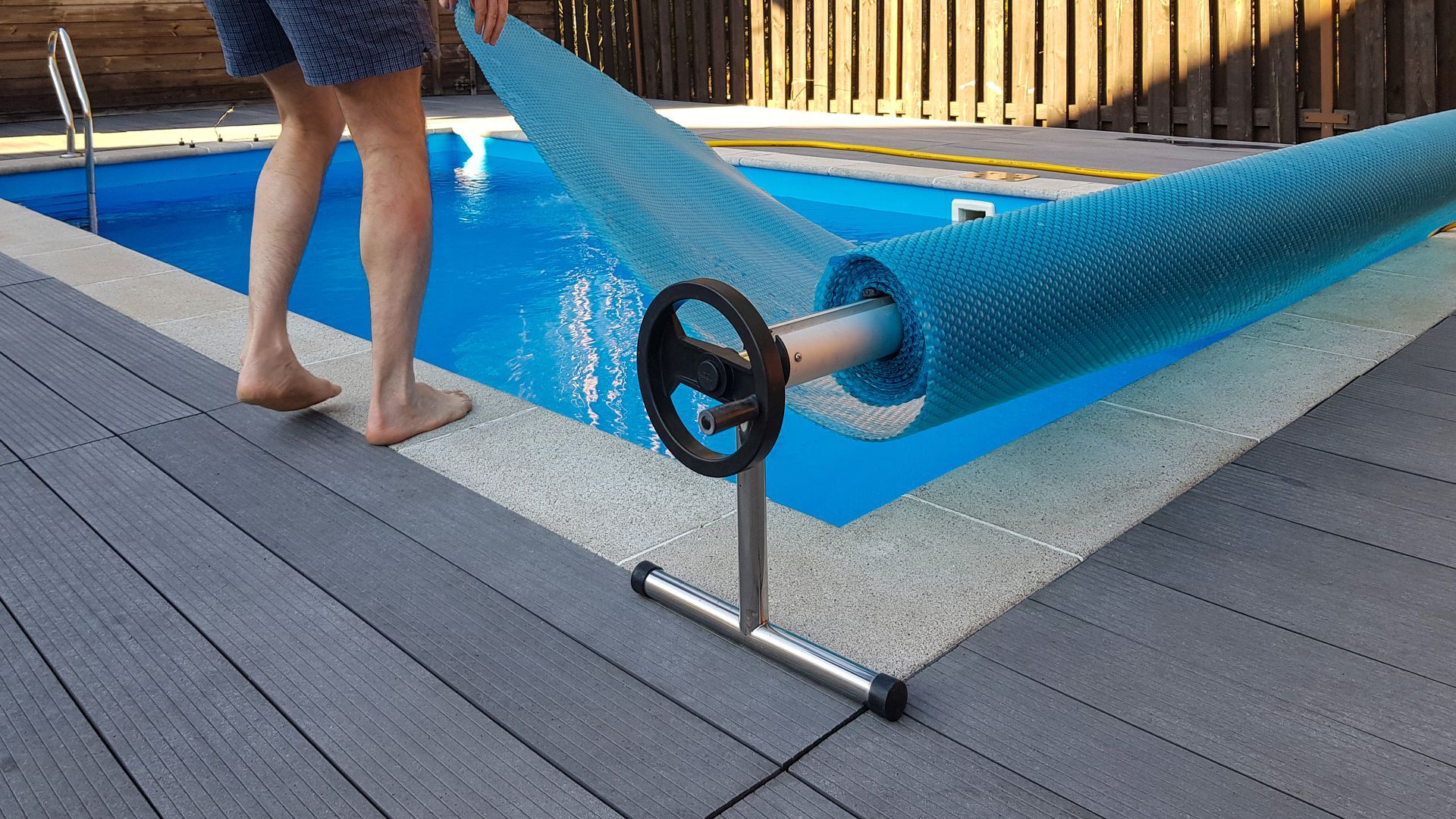 Une personne déplie une bâche bleue sur une piscine hors sol sur une terrasse grise.