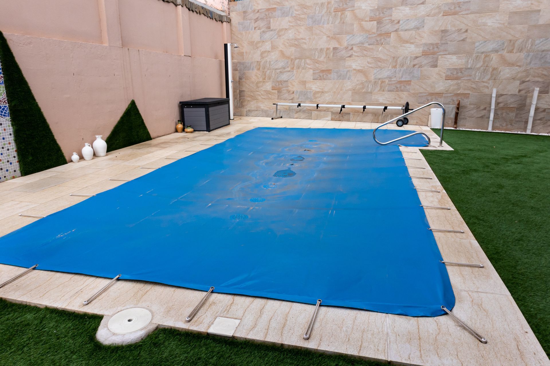 Piscine de jardin recouverte d'une bâche bleue, jouxtant une pelouse artificielle et un mur beige.