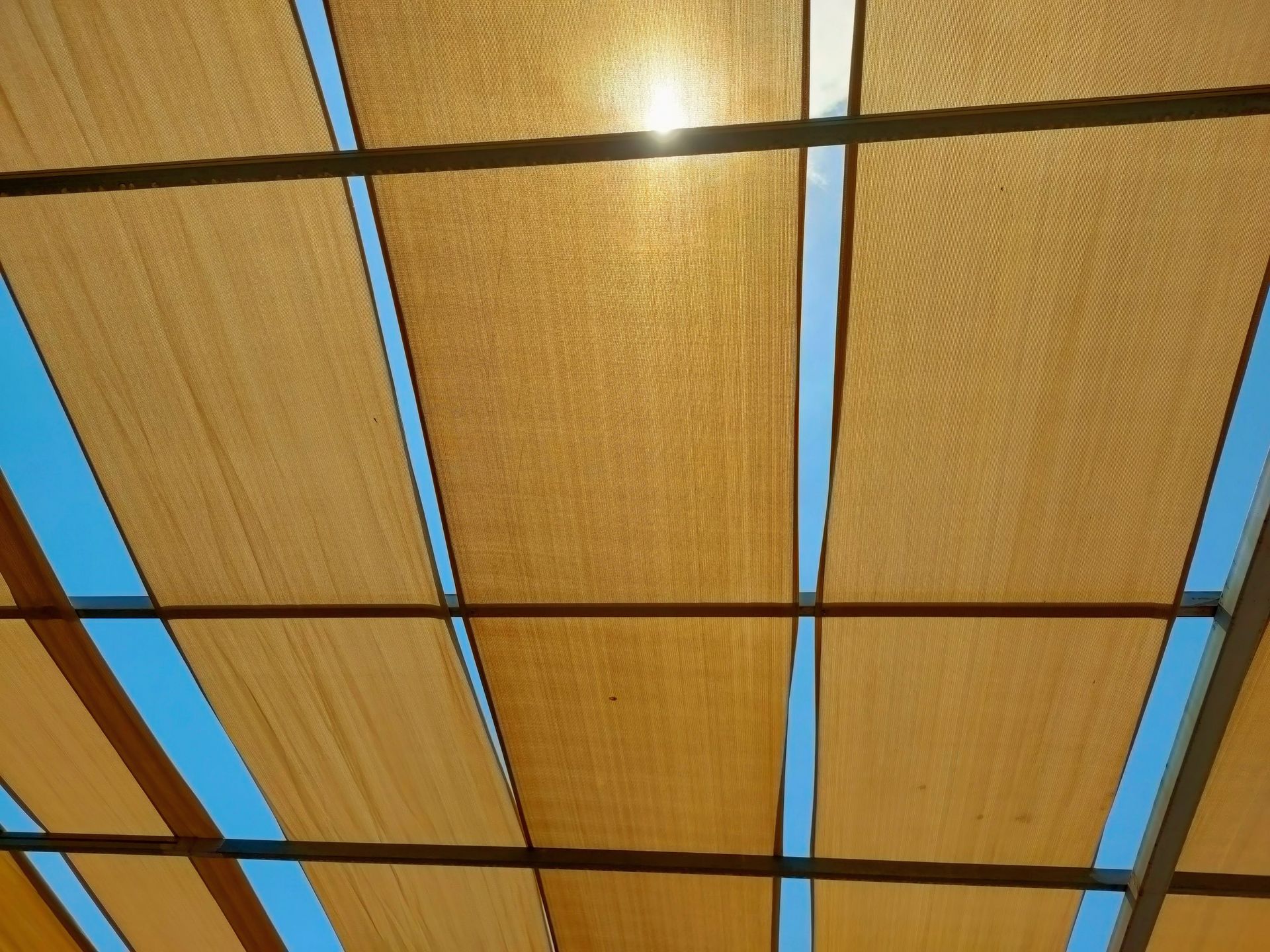 La lumière du soleil traverse des toiles beiges et laisse entrevoir le ciel bleu entre les poutres sombres de la structure.