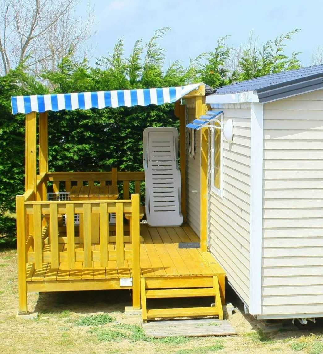 Terrasse jaune avec un auvent à rayures bleues et blanches à côté d'un mobile home et de buissons verts.