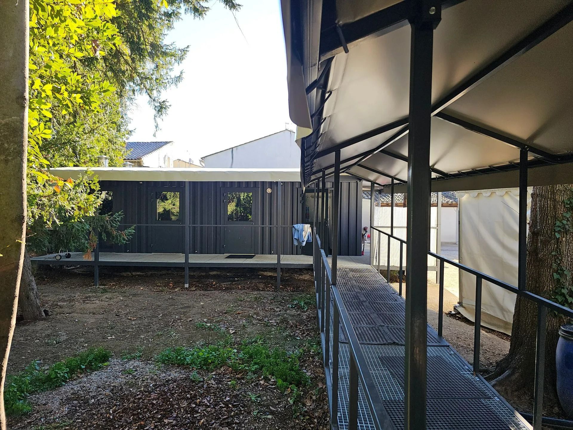 Allée extérieure longeant une rangée de tentes et un petit bâtiment, avec des arbres et un terrain en terre à proximité.