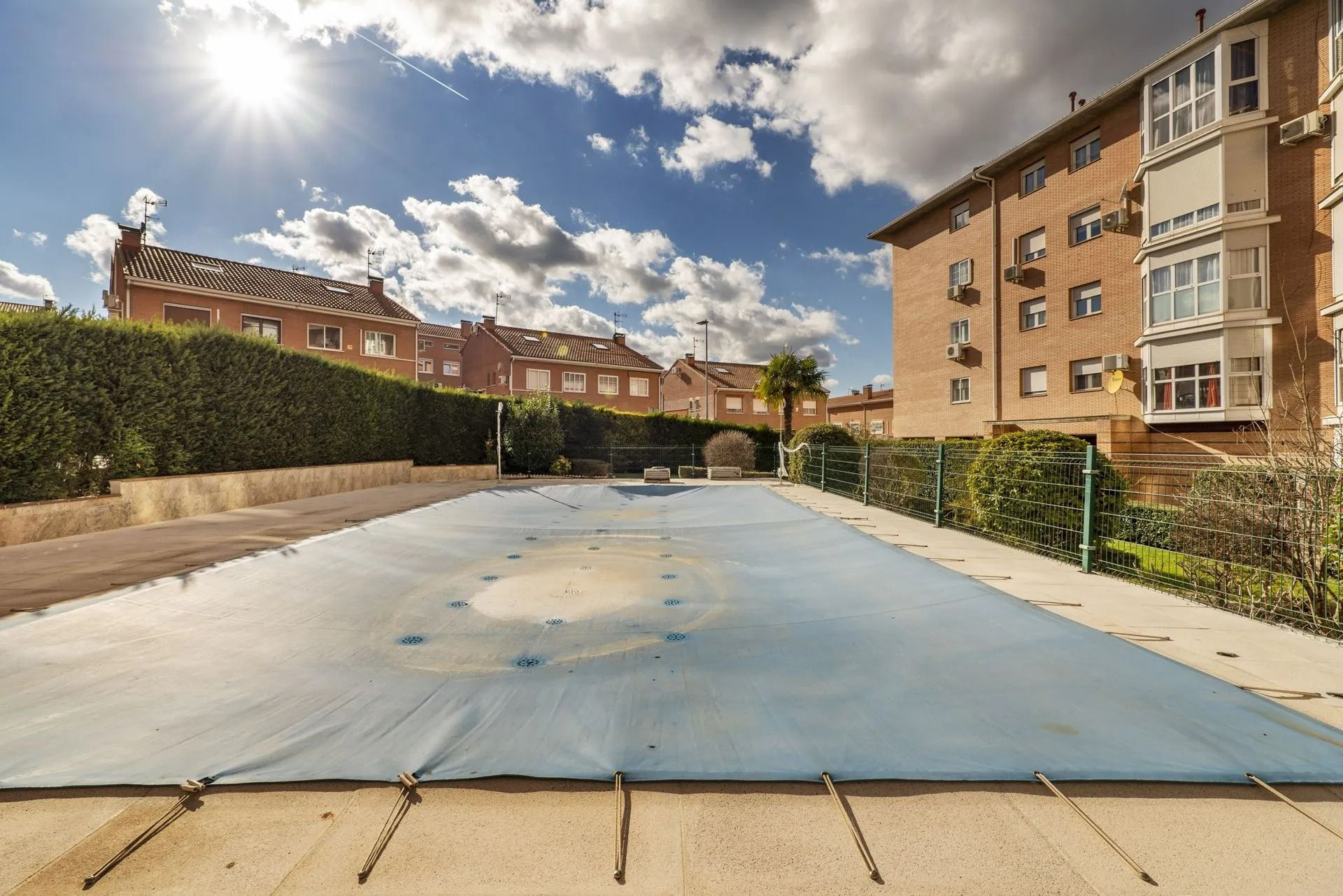 Cour extérieure vide avec piscine couverte d'une bâche bleue, entourée d'immeubles et de haies sous un ciel ensoleillé