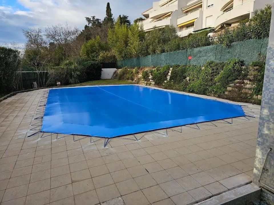 Piscine couverte d'une bâche bleue sur une terrasse carrelée et des haies dans une cour résidentielle