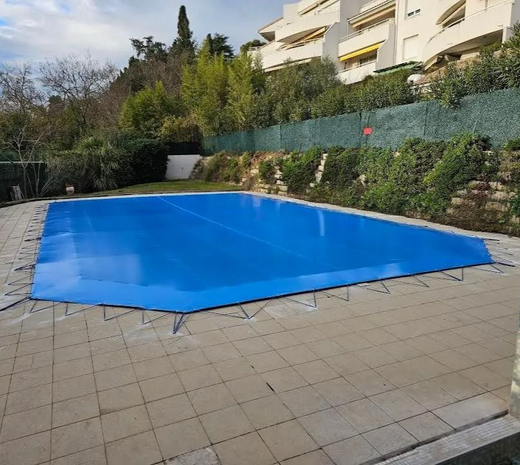 Piscine recouverte d'une bâche bleue dans une cour extérieure pavée, avec des arbustes et des immeubles à proximité.