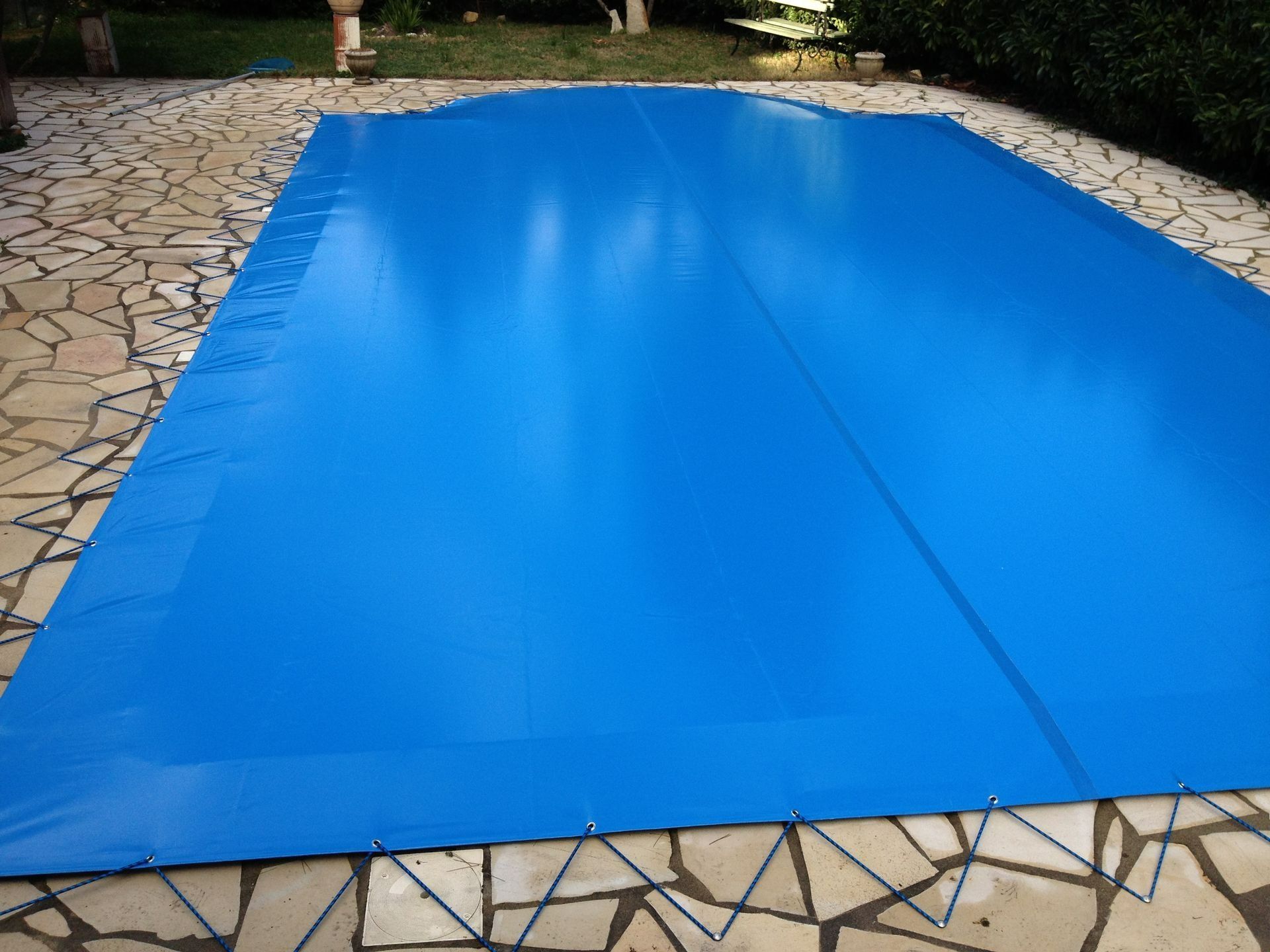 Une bâche bleue recouvre une piscine rectangulaire dans une cour carrelée