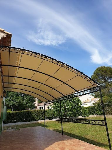 Terrasse extérieure couverte avec un auvent beige, une structure métallique et une pelouse verte sous un ciel bleu.