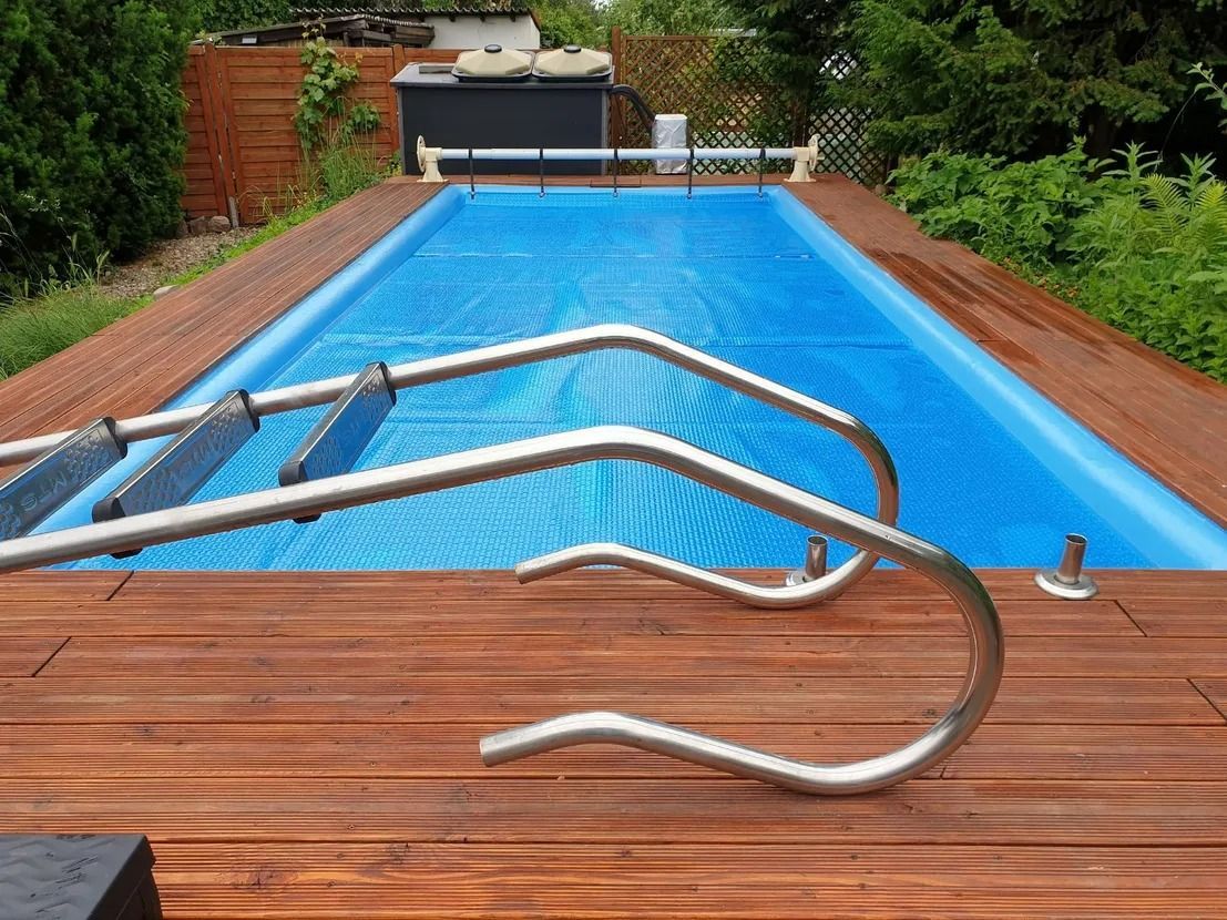 Piscine extérieure couverte avec une bâche bleue et une échelle de piscine posée sur la terrasse en bois