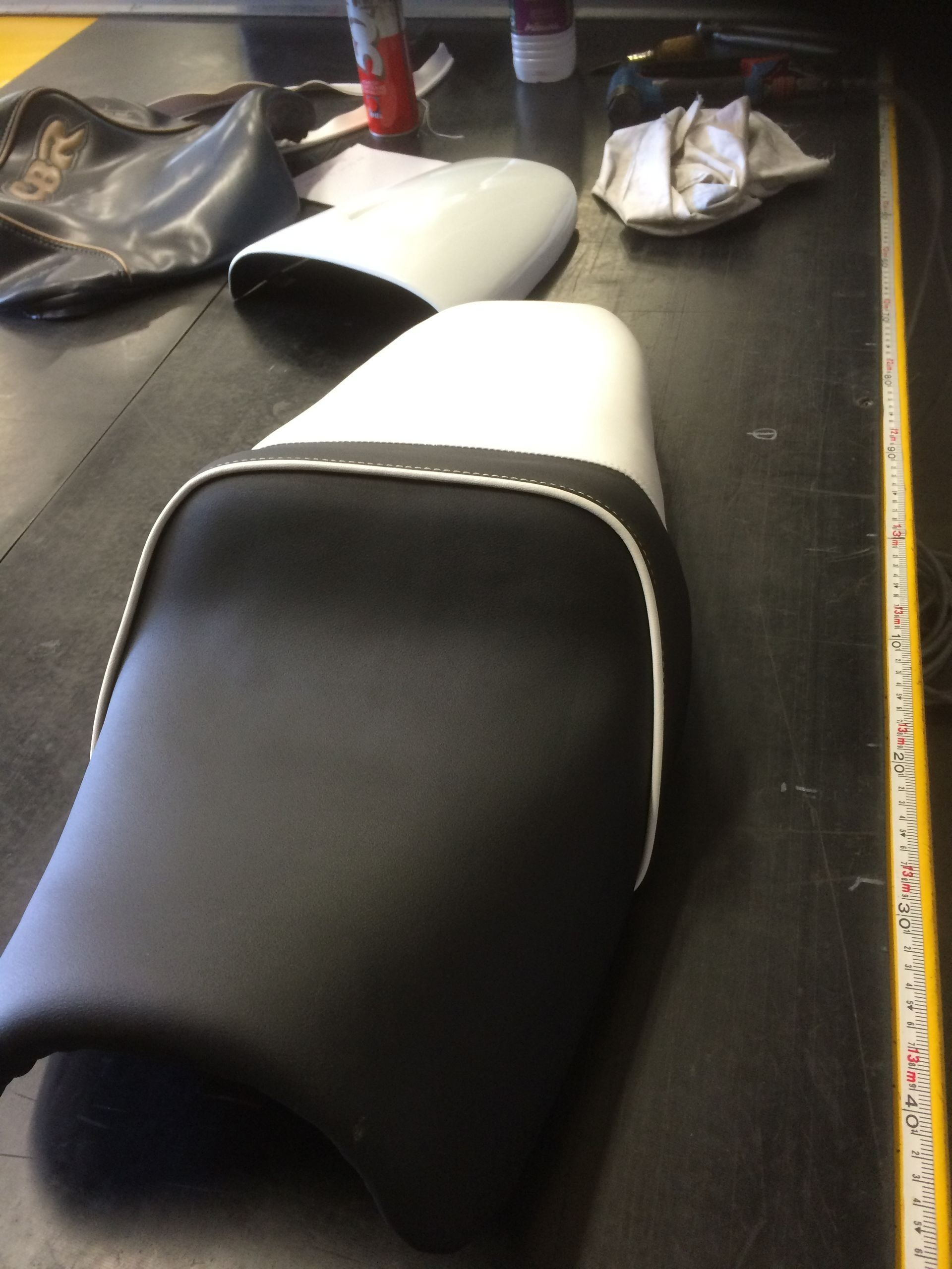 Des housses de selle de moto noires et blanches sur un établi, avec des outils et un mètre ruban.