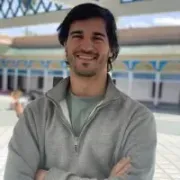 Un hombre con barba sonríe con los brazos cruzados frente a un edificio.