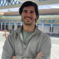 Un hombre con barba sonríe con los brazos cruzados frente a un edificio.