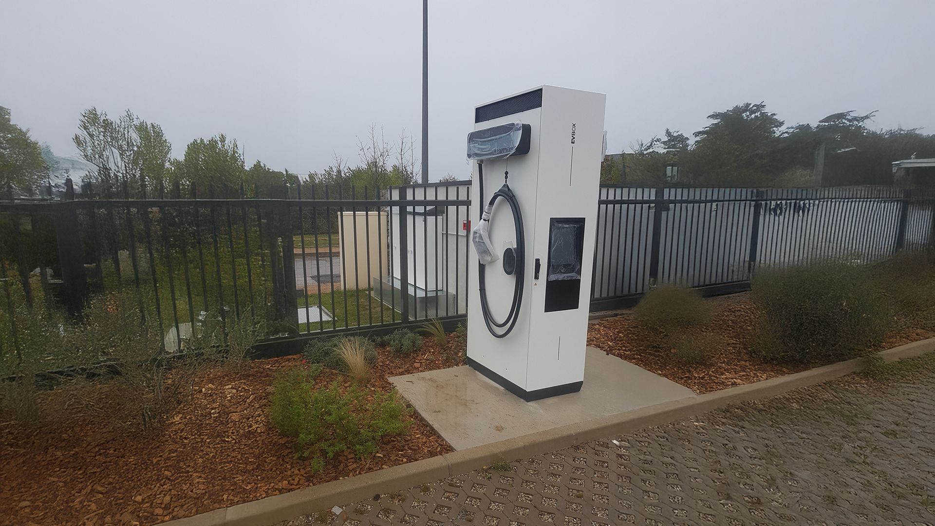 Borne de recharge pour véhicules électriques sur une dalle de béton, entourée d'une clôture noire et de verdure. Ciel couvert.