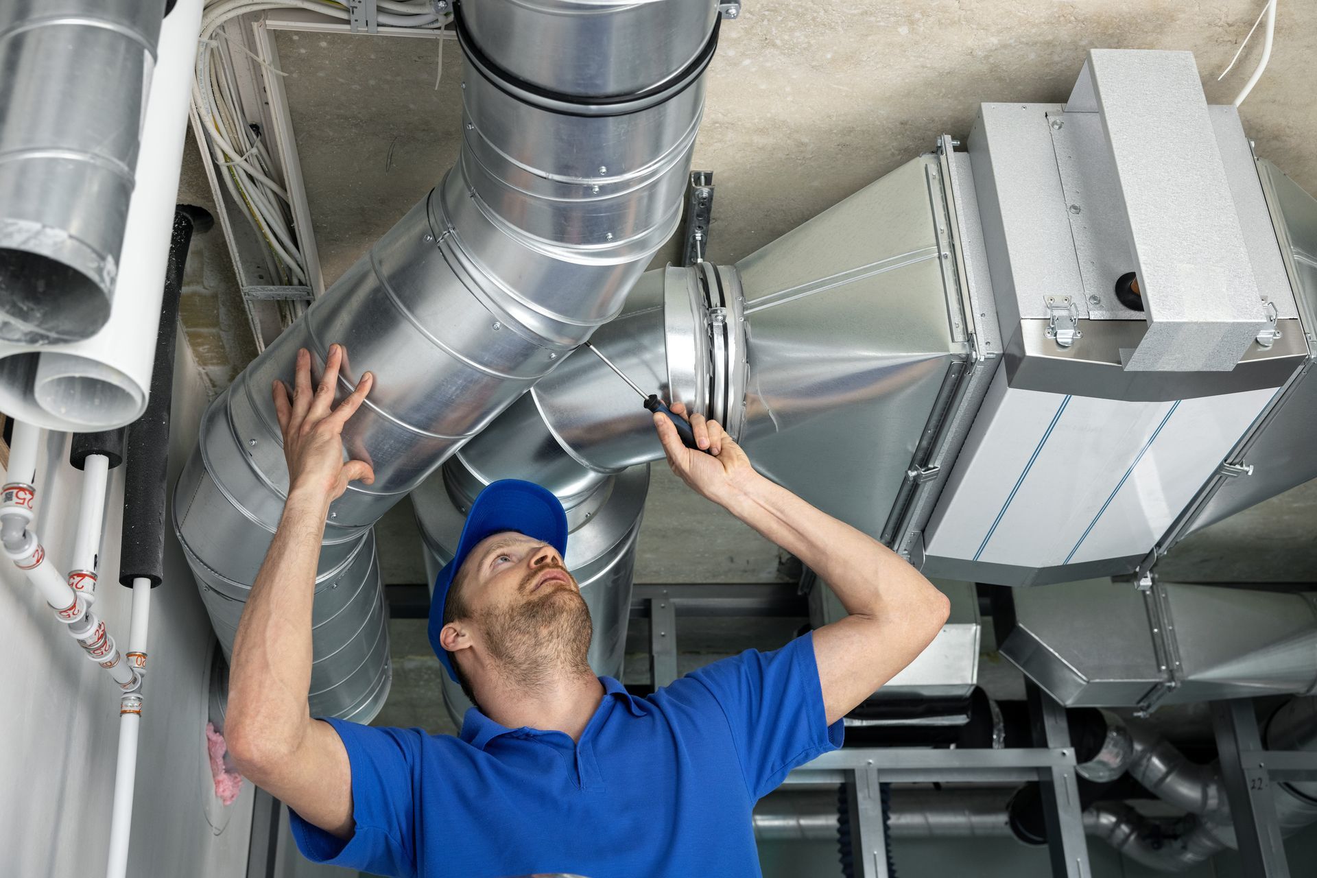 Un technicien en chemise bleue inspecte des conduits de ventilation au plafond.