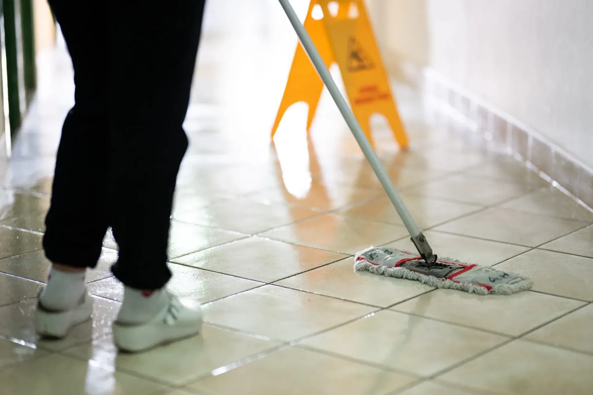 Persona trapeando un piso de baldosas, señal de precaución amarilla en el fondo.