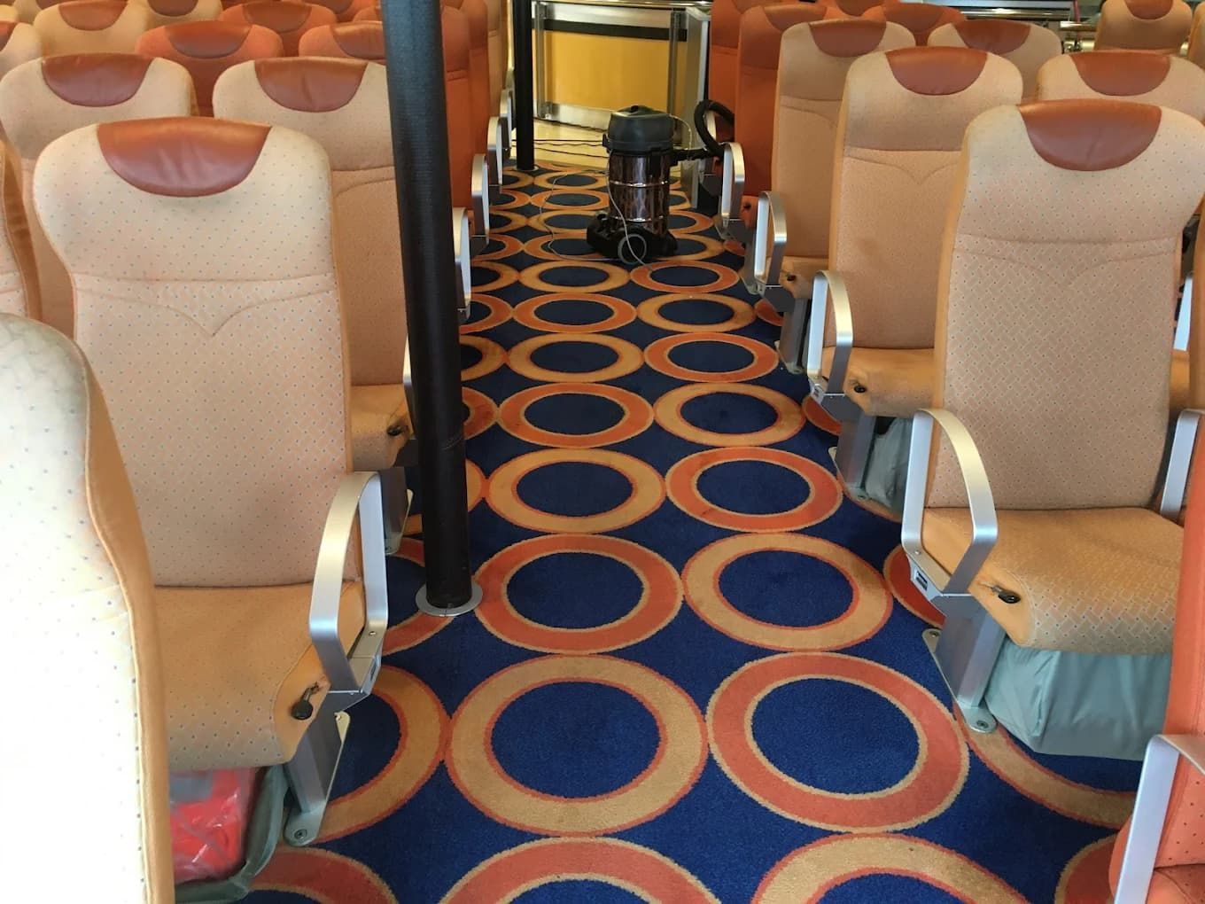 Interior de un autobús, filas de asientos color melocotón con apoyabrazos de metal, alfombra estampada en azul y naranja.
