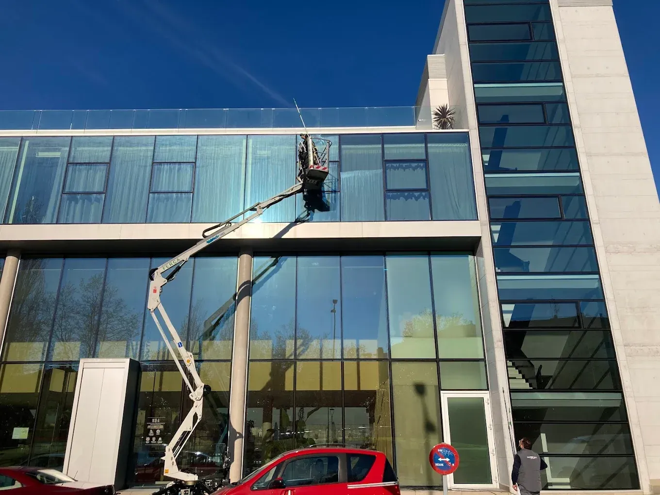 Persona en un ascensor limpiando ventanas de un edificio moderno con vidrios tintados de azul en un día soleado.