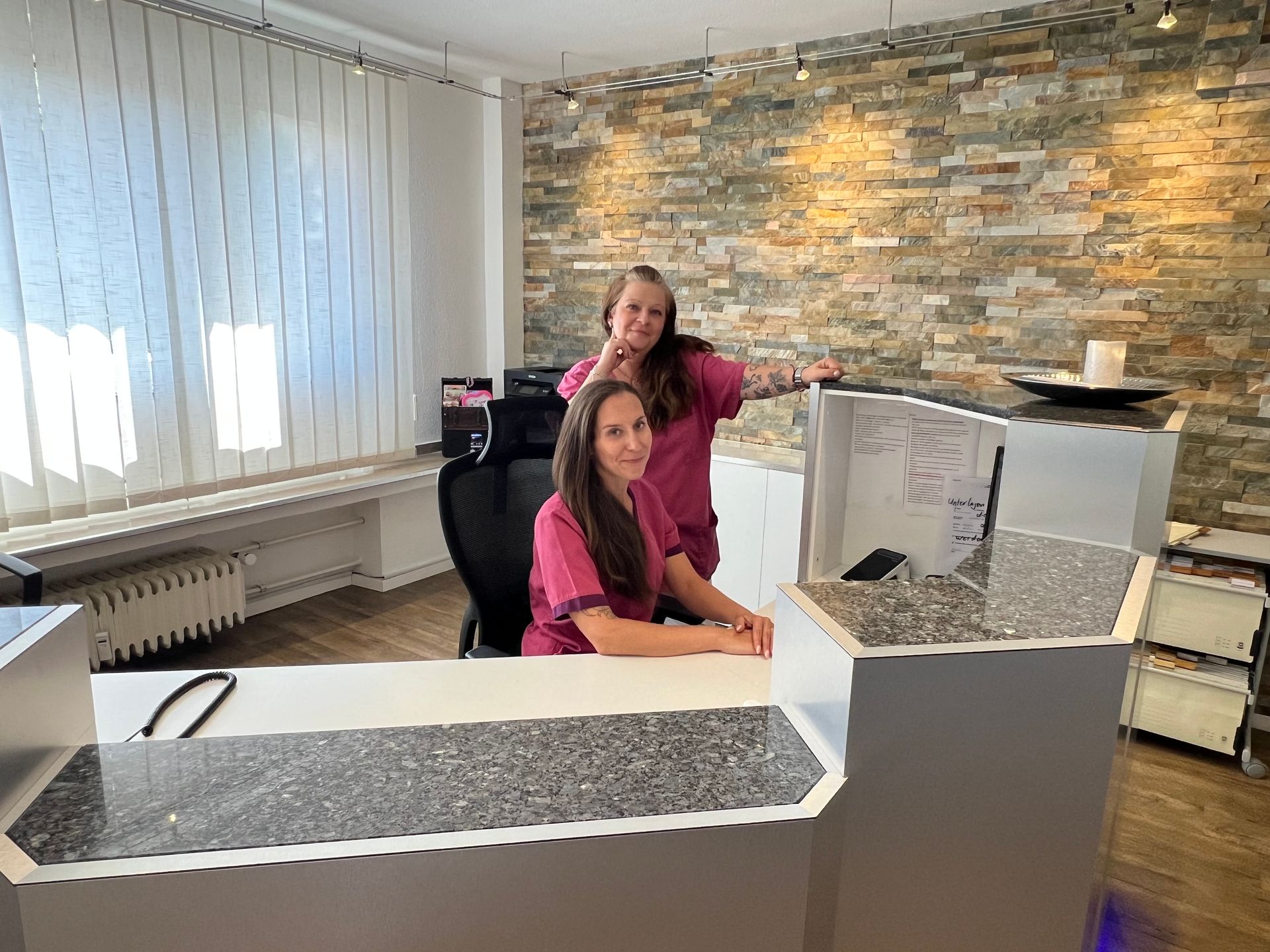 Zwei Frauen in Pink hinter einem weißen Empfangstresen in einem hellen Büro mit einer Akzentwand aus Stein.