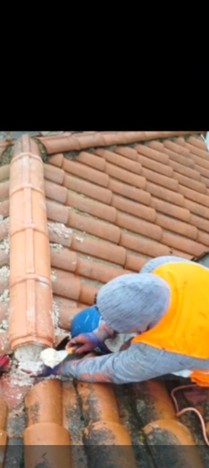 Trabajador con chaleco naranja reparando tejas de arcilla en un tejado inclinado.