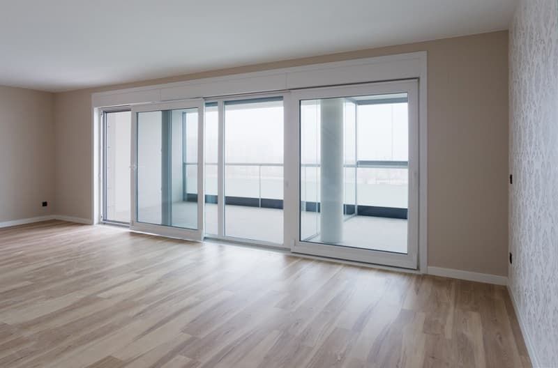 Salón moderno y vacío con suelos de madera clara y grandes puertas correderas de cristal que dan a un balcón.