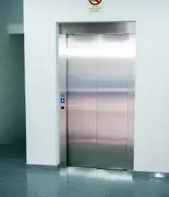 Vista interna de um elevador com paredes brancas, piso cinza, portas fechadas e painel de controle.