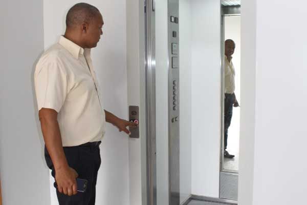 Elevador externo de vidro em um prédio com janelas de tonalidade verde contra um céu azul.