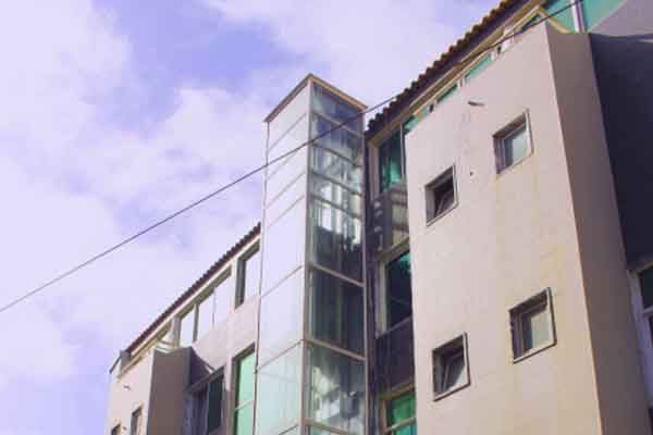 Elevador de vidro na fachada de um prédio de vários andares. Céu nublado.