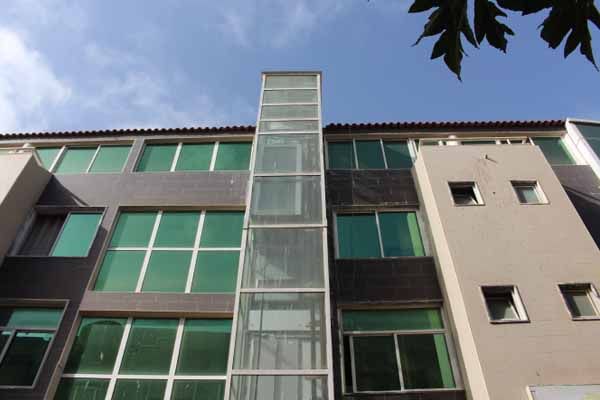 Elevador externo de vidro em um prédio com janelas de tonalidade verde contra um céu azul.