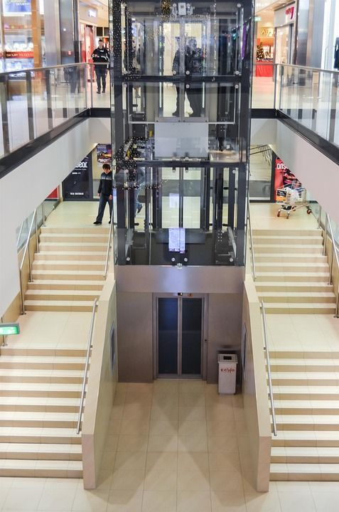 Elevador em um shopping center. Escadas em ambos os lados. Pessoas visíveis nos andares superiores.