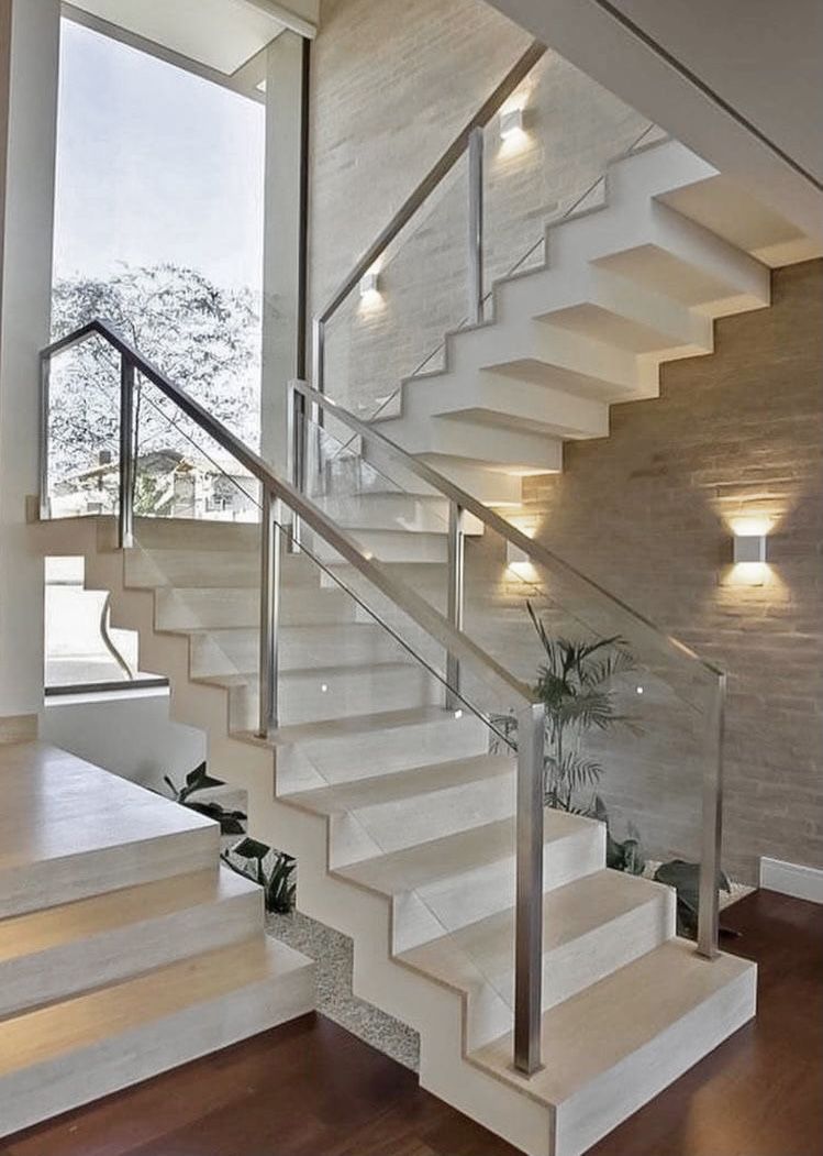 Escada moderna em tons claros com corrimão de vidro e metal. Iluminada por apliques de parede.