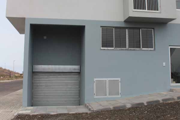 Prédio cinza com porta de enrolar, janelas e entrada. Guia de concreto.