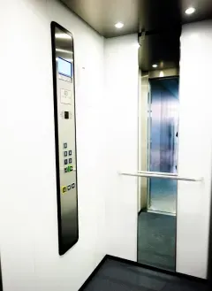 Interior de elevador com portas prateadas fechadas, indicador de andar e luzes brilhantes.