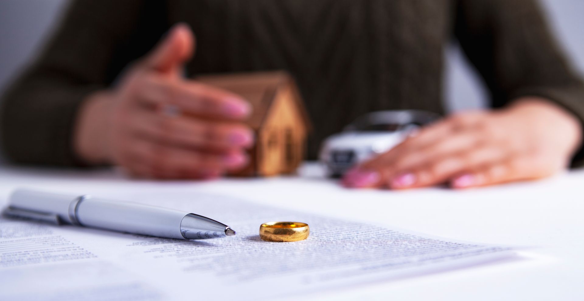 Une bague en or posée sur un document. À l'arrière-plan, une personne tient une maquette de maison et une voiture miniature.