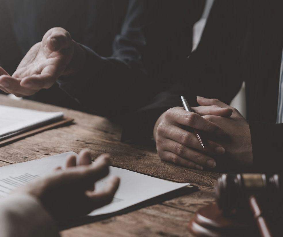 Trois personnes discutent à table. L'un d'entre eux tient un stylo. Des documents sont visibles.