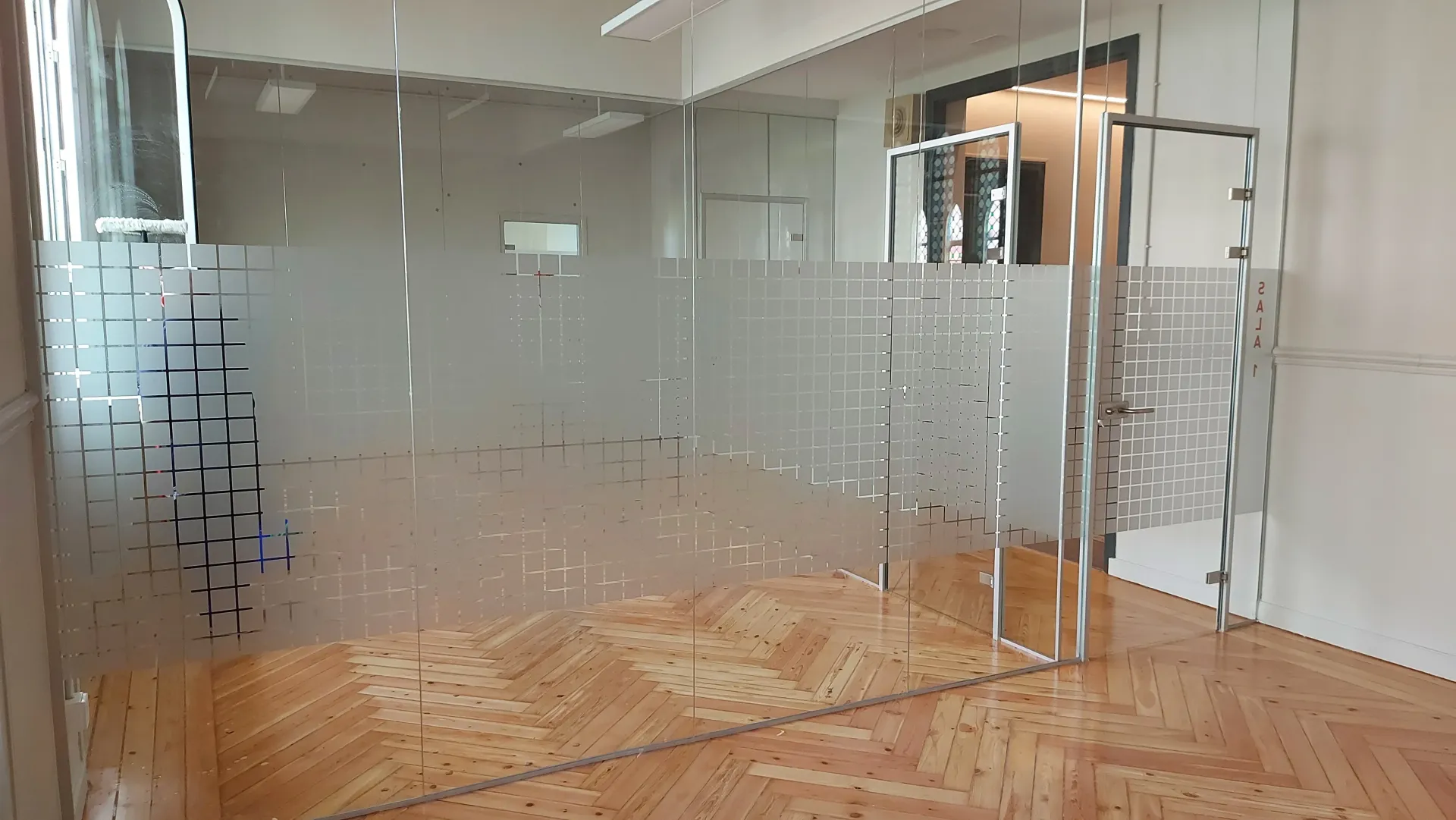Oficina moderna con paredes de cristal, paneles esmerilados, suelo de madera y puerta de cristal abierta.