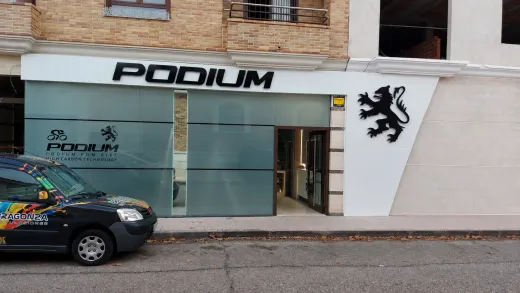 Fachada con letrero “PODIUM”, entrada de cristal, logotipo de león negro y un coche aparcado en el exterior.