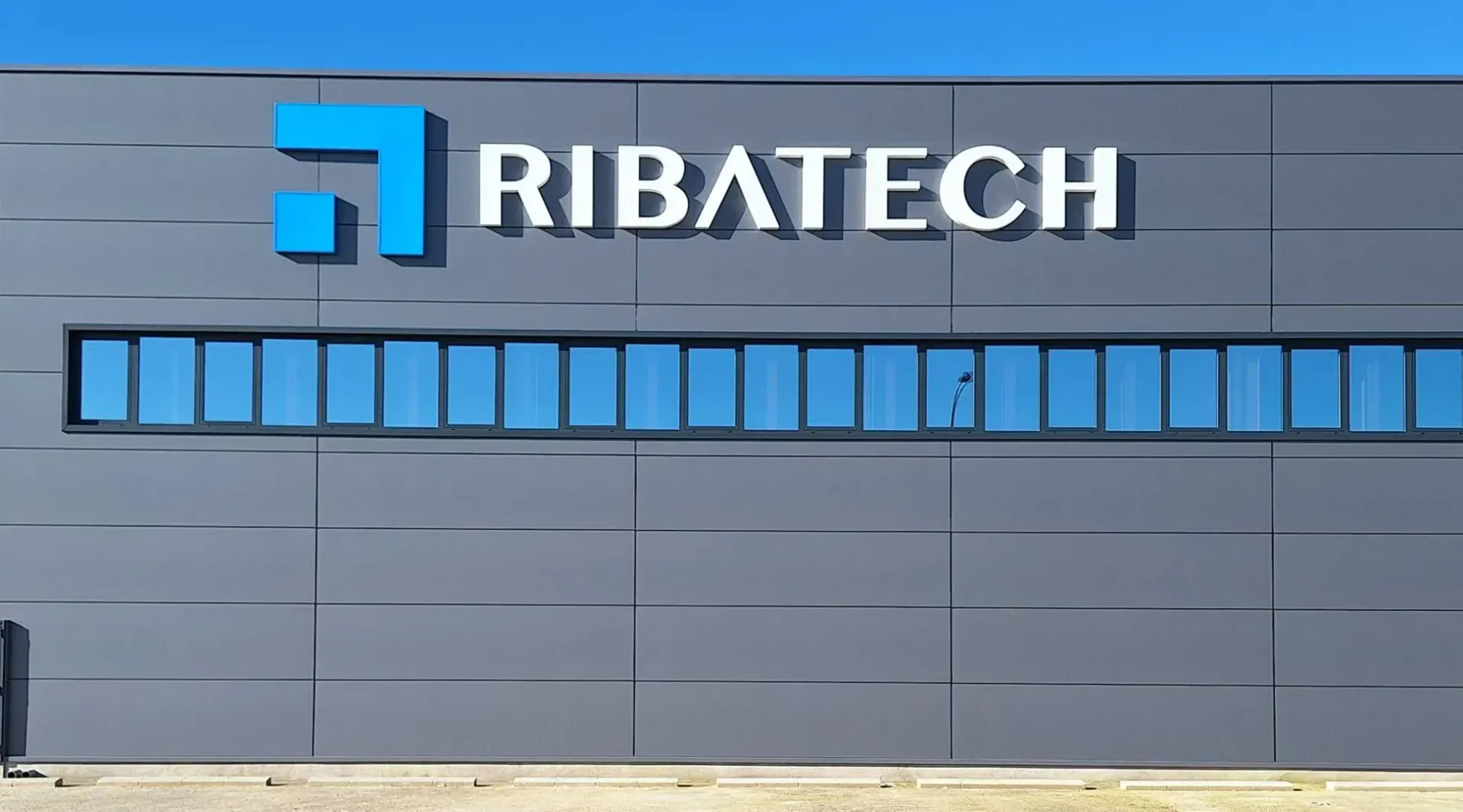 Letrero de RIBATECH en un edificio gris con ventanas azules y logotipo azul.