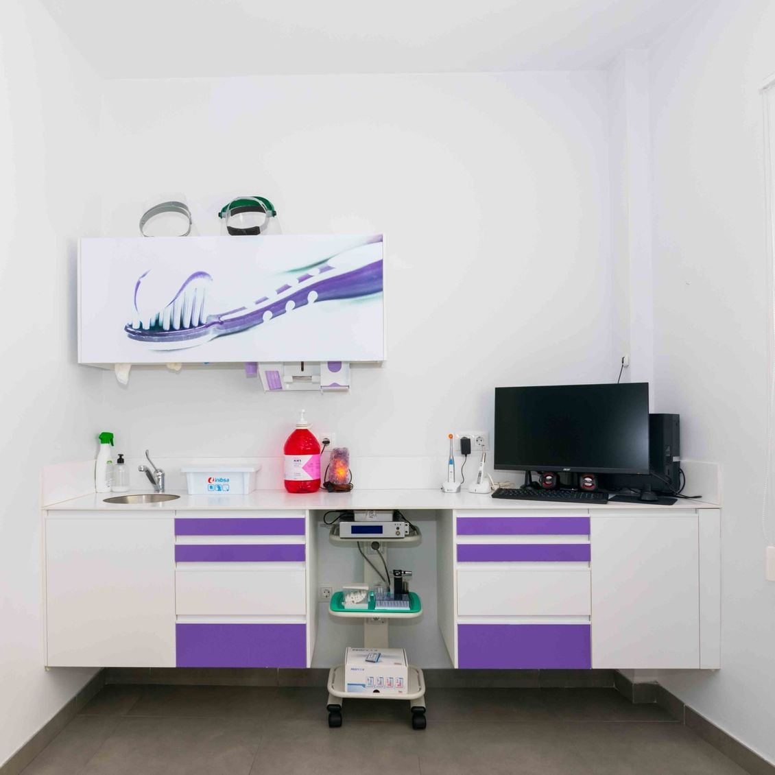 Un consultorio dental con gabinetes blancos y cajones morados y una computadora en el escritorio.