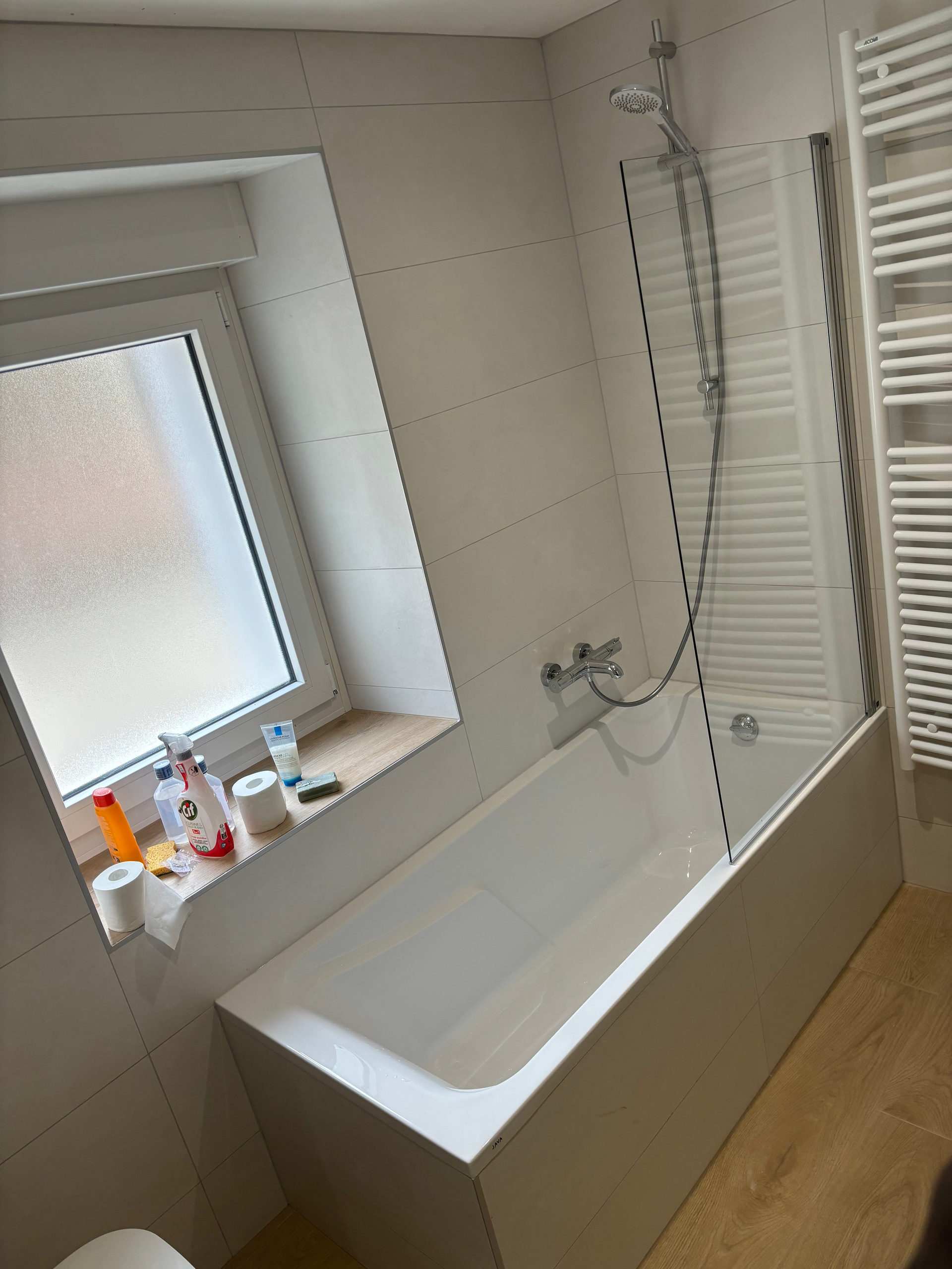 Petite salle de bains blanche avec baignoire, murs carrelés, étagère sous la fenêtre et pommeau de douche sur la droite.