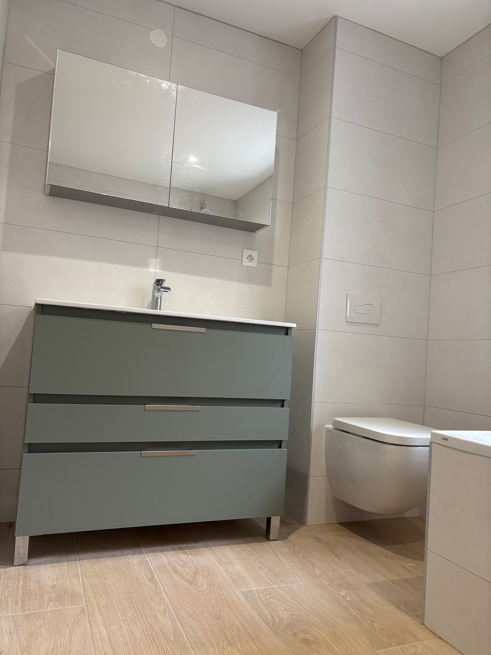 Petite salle de bains moderne avec meuble-lavabo gris, miroir, lavabo et toilettes sur un sol imitation bois.