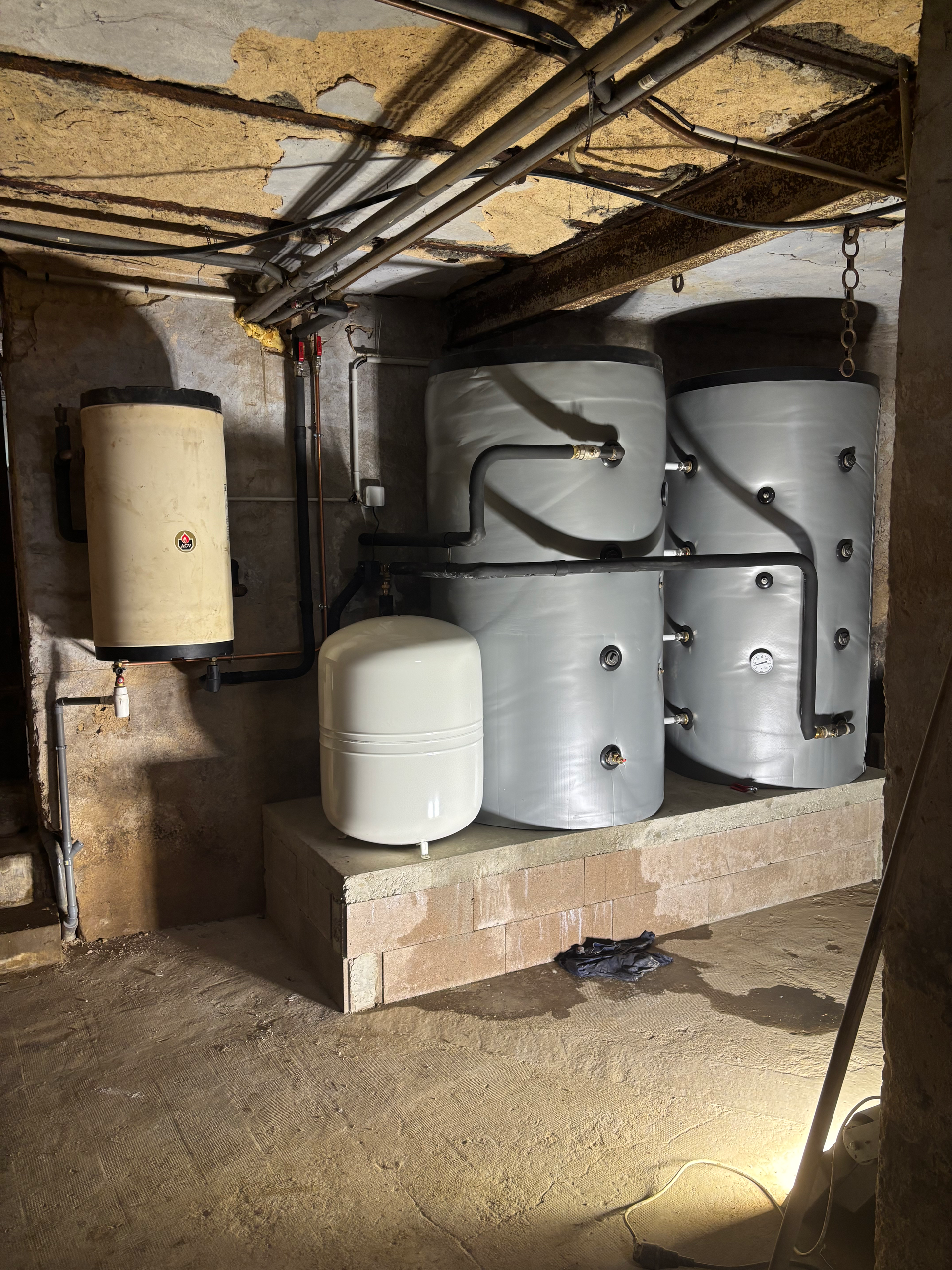 Local technique avec plusieurs chauffe-eau et réservoirs sous un plafond bas, tuyauterie apparente et sol en béton.