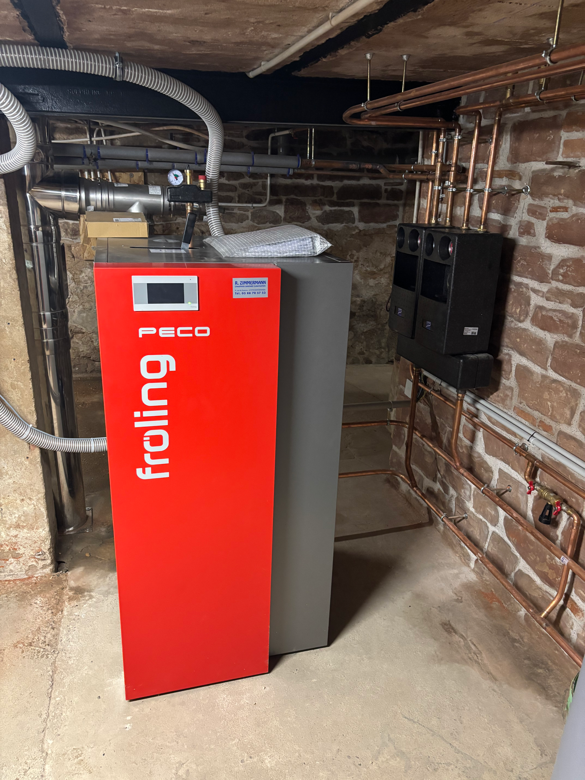Chaudière rouge dans un sous-sol en briques, à côté d'un appareil électroménager gris et de tuyaux apparents sous le plafond.