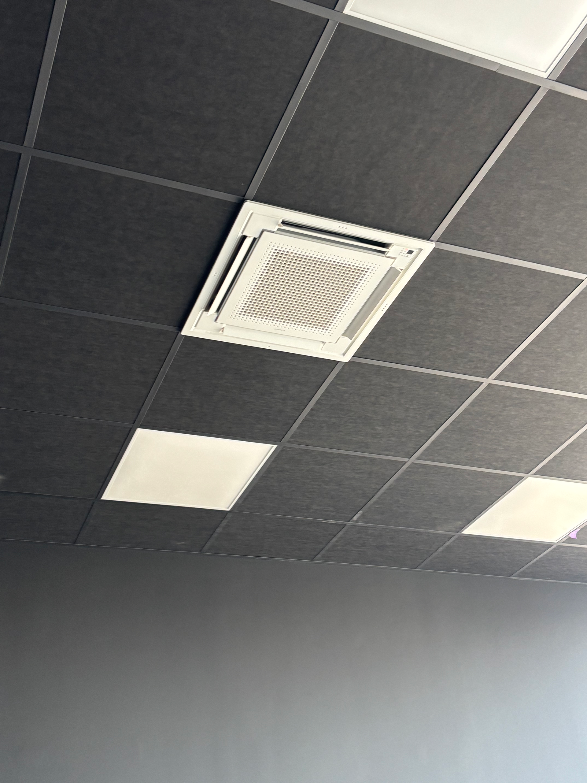 Plafond de bureau avec dalles acoustiques noires, une bouche d'aération centrale et deux panneaux lumineux blancs.