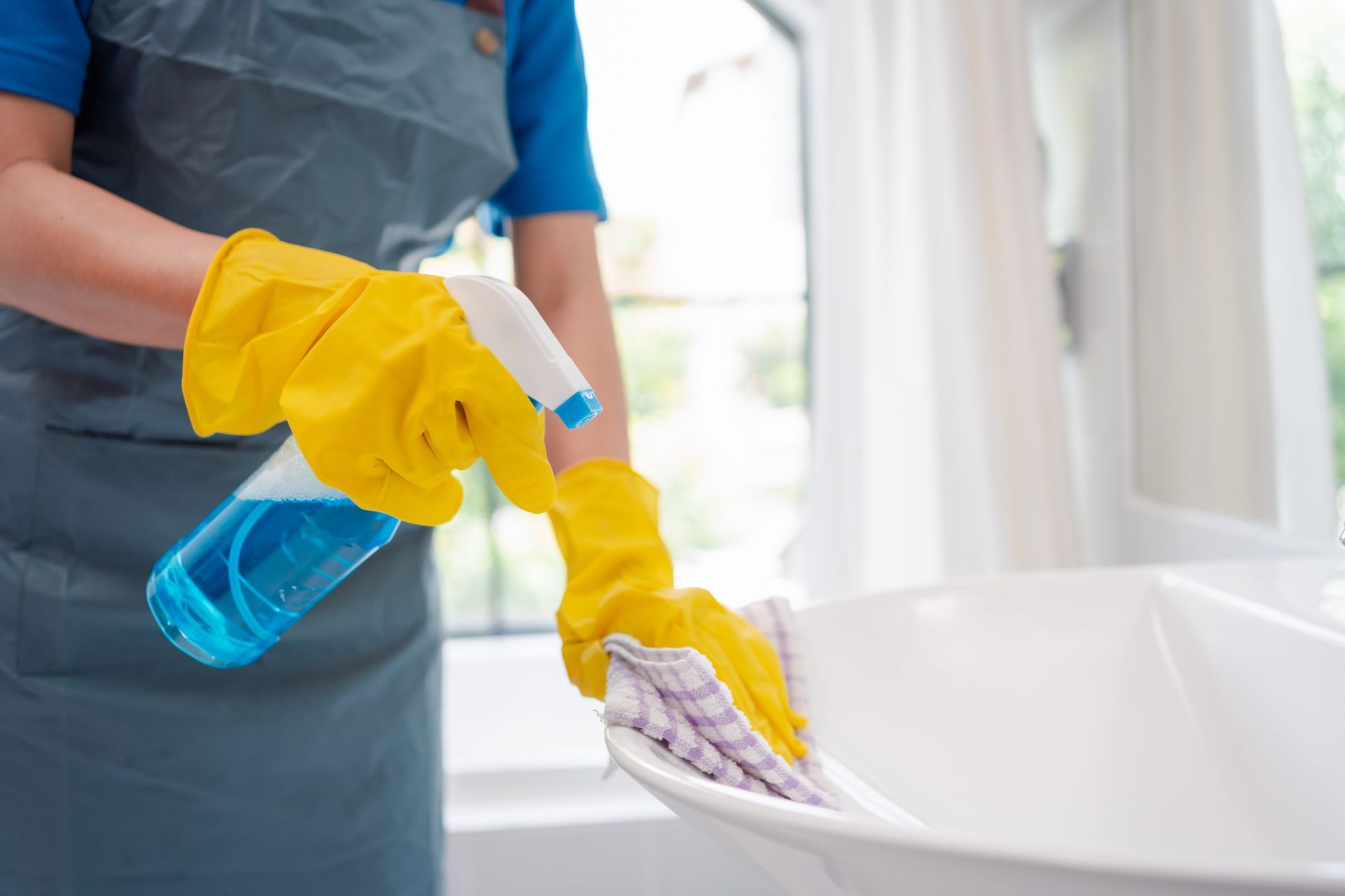 Une personne portant des gants jaunes vaporise et essuie une baignoire blanche avec un produit nettoyant bleu.