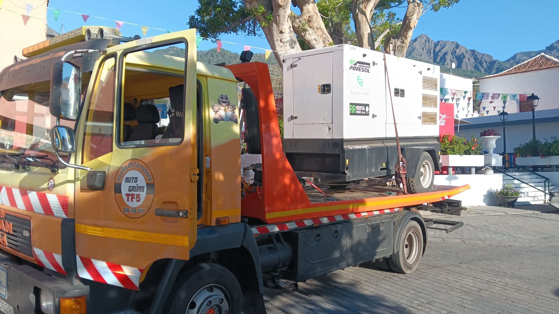 Una grúa que transporta una retroexcavadora amarilla en una calle de la ciudad junto a un edificio de color beige.