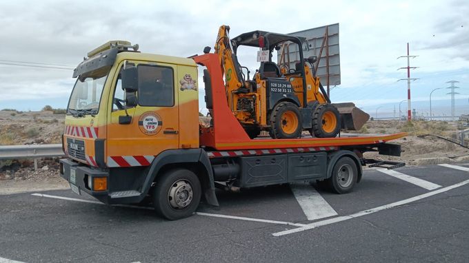 Una miniexcavadora amarilla sobre un camión de plataforma está estacionada junto a un edificio verde y gris.