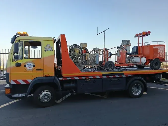 Grúa de plataforma amarilla transportando maquinaria industrial naranja en una carretera bajo un cielo despejado.